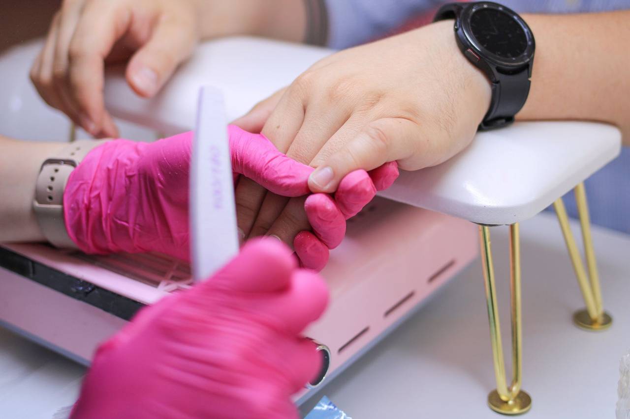 A technician in pink gloves shaping a client’s nails